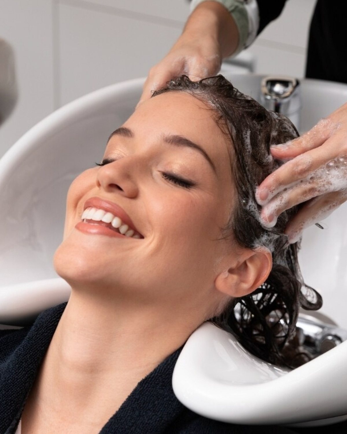 Smiling woman relaxing at the hair wash basin while a stylist massages her scalp during the Advanced Scalp Detox at Luxe Elegance Beauty Center