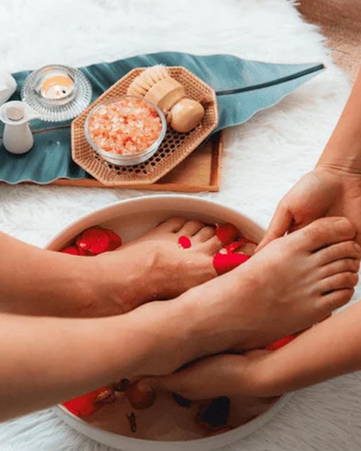Therapist gently washing a client's feet in a bowl of warm water with red rose petals, beside a spa tray with a candle, bath salts and a soft towel