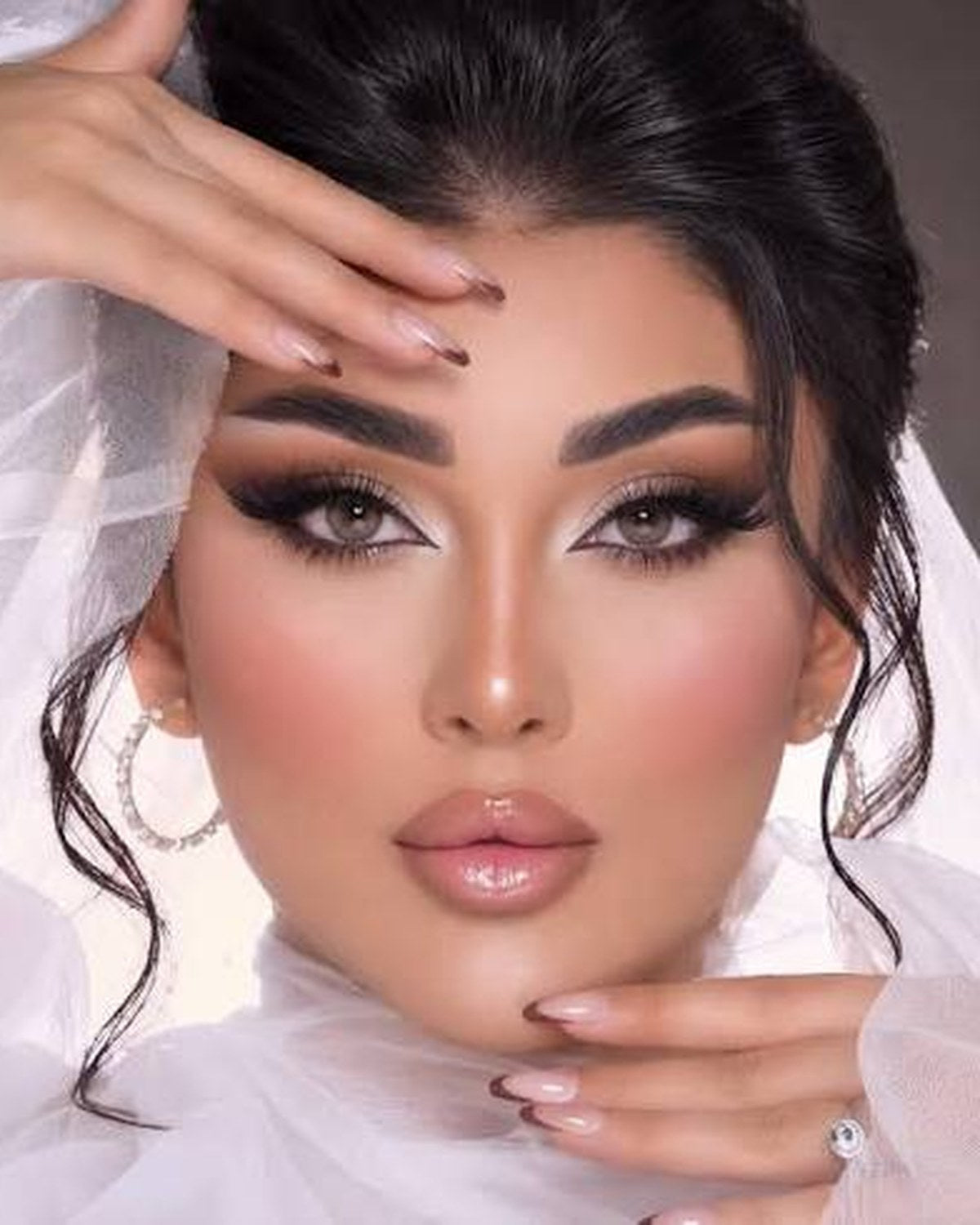 Full glam bridal makeup look with soft brown smoky eyes, winged lashes, sculpted glowing skin and glossy nude lips, with dark hair in a soft updo and a white veil