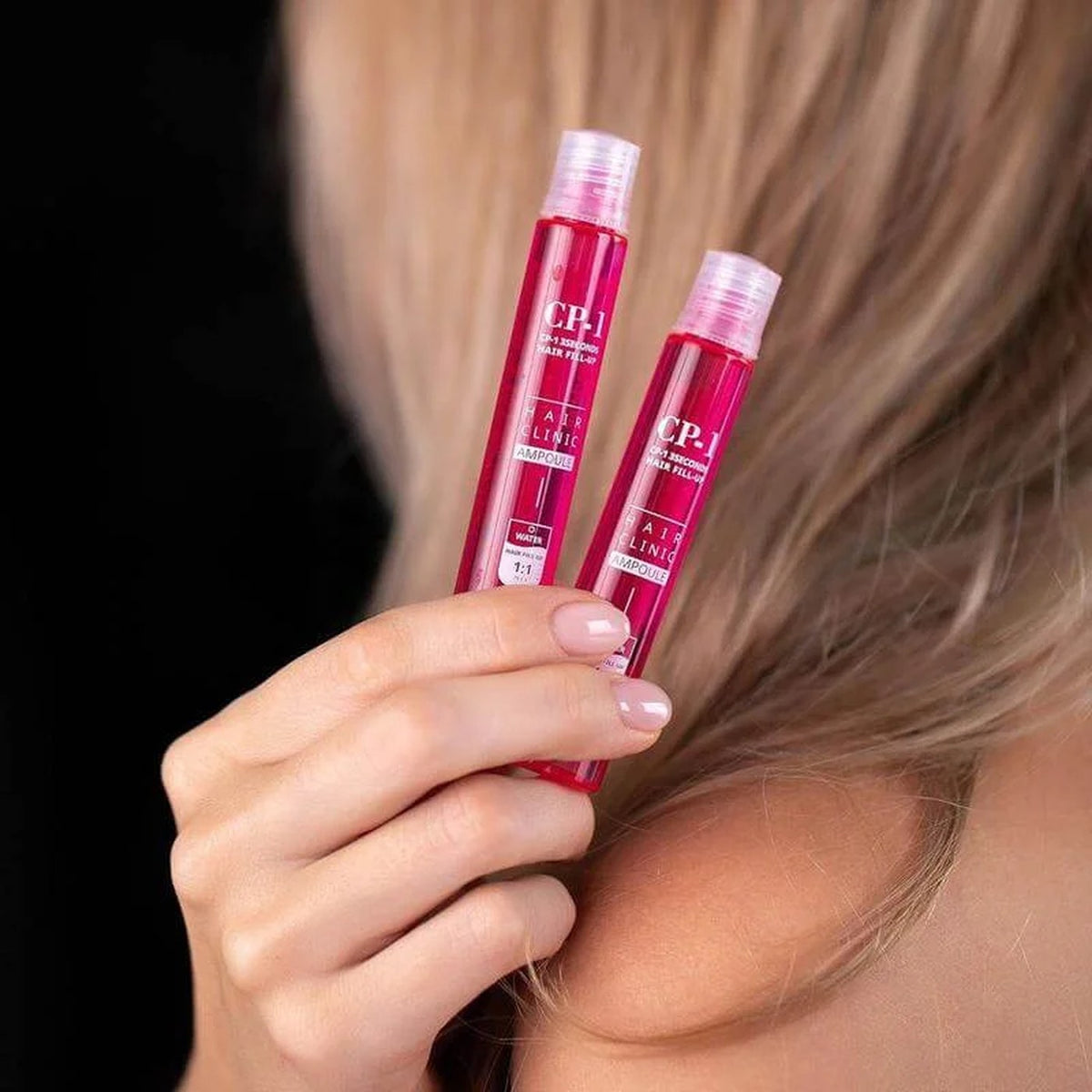 Hand holding two pink hair filler ampoules next to blonde hair, the hair filler ampoule option of the Hair Glow treatment