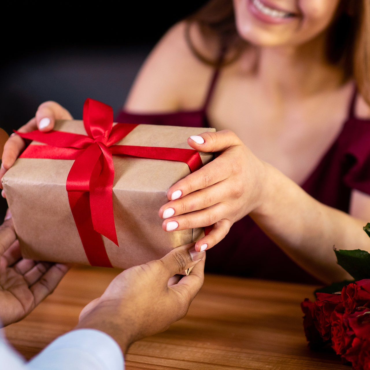 Person receiving a gift wrapped in brown paper with a red ribbon from another person, with a glass of red wine and flowers in the background.