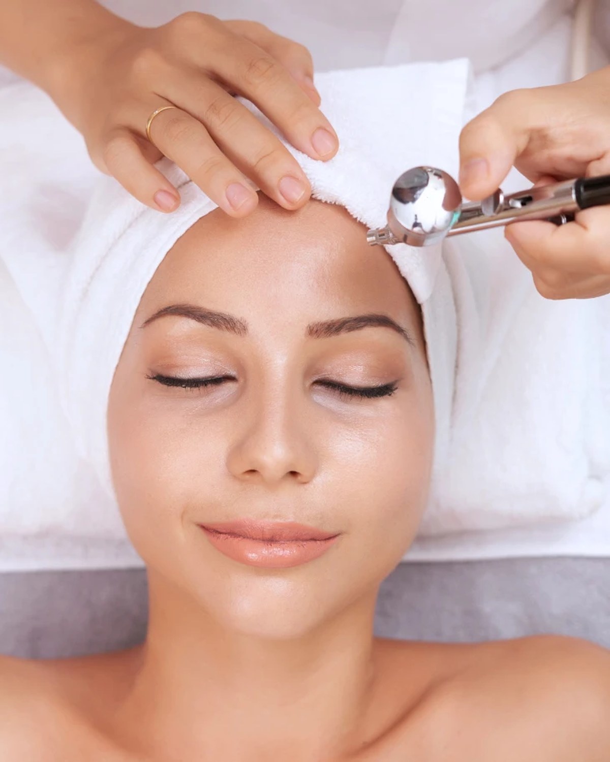 Woman relaxing with eyes closed and hair wrapped in a white towel while a beautician sprays oxygen onto her skin during an Oxygen Radiance facial