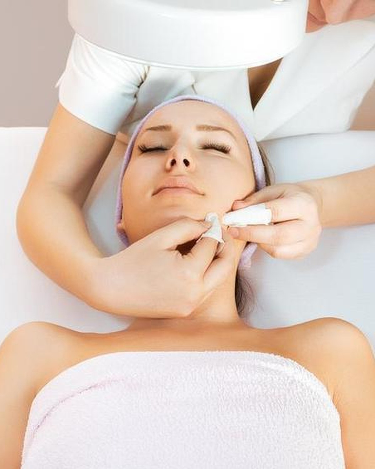 Woman relaxing with eyes closed on a treatment bed while a beautician gently cleanses her skin during a signature deep cleansing facial