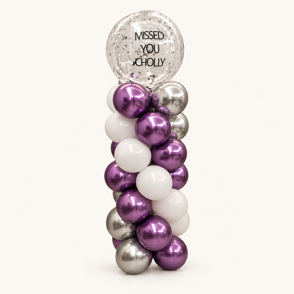 Purple, white and silver spiral balloon column topped with a clear confetti balloon reading Missed You Cholly.
