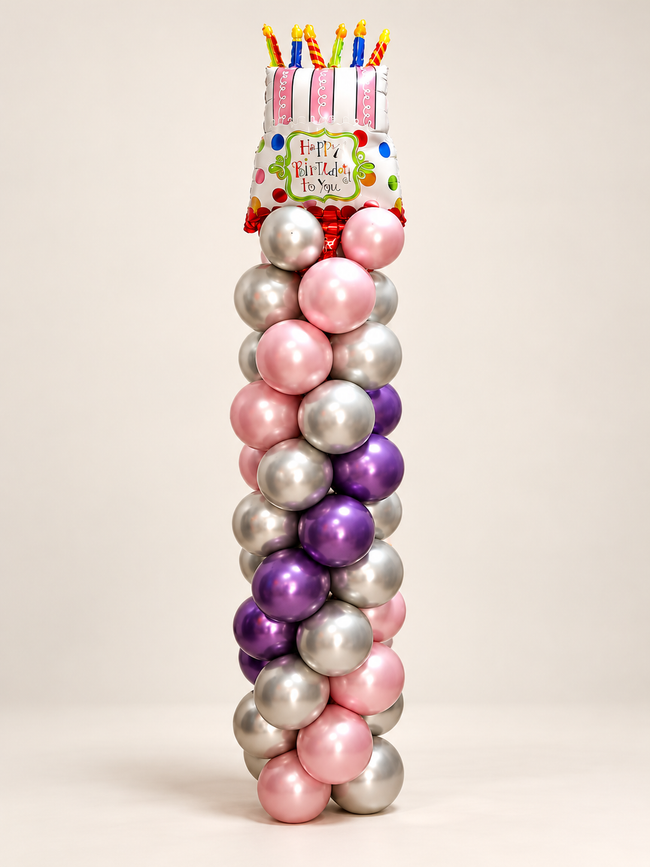 Birthday Royale metallic pink, purple and silver balloon column topped with a birthday cake-shaped foil balloon and colorful candle shapes on an off-white background