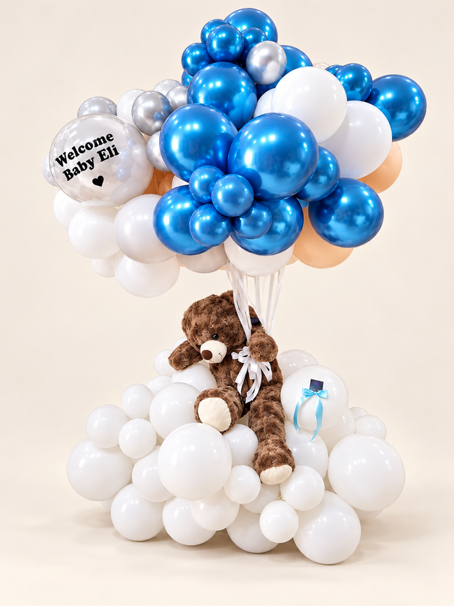 Grand Teddy Welcome with blue, silver, white and peach balloons, a personalized Welcome Baby Eli clear bubble, large brown teddy and white balloon base