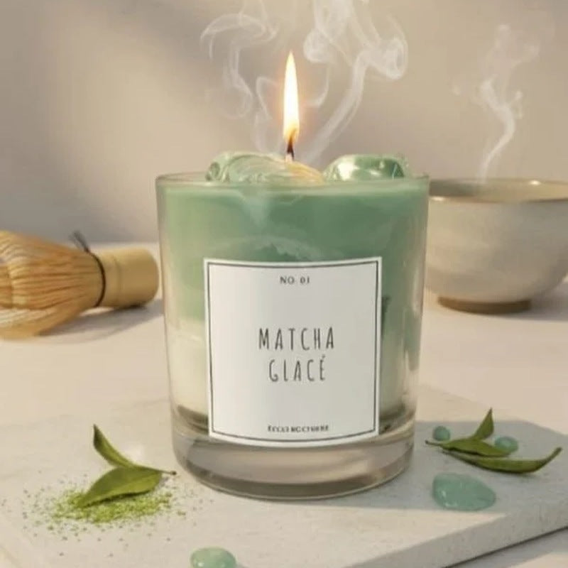 Lit matcha-scented candle in a glass jar with a white label, beside a bamboo whisk, tea leaves and green stones