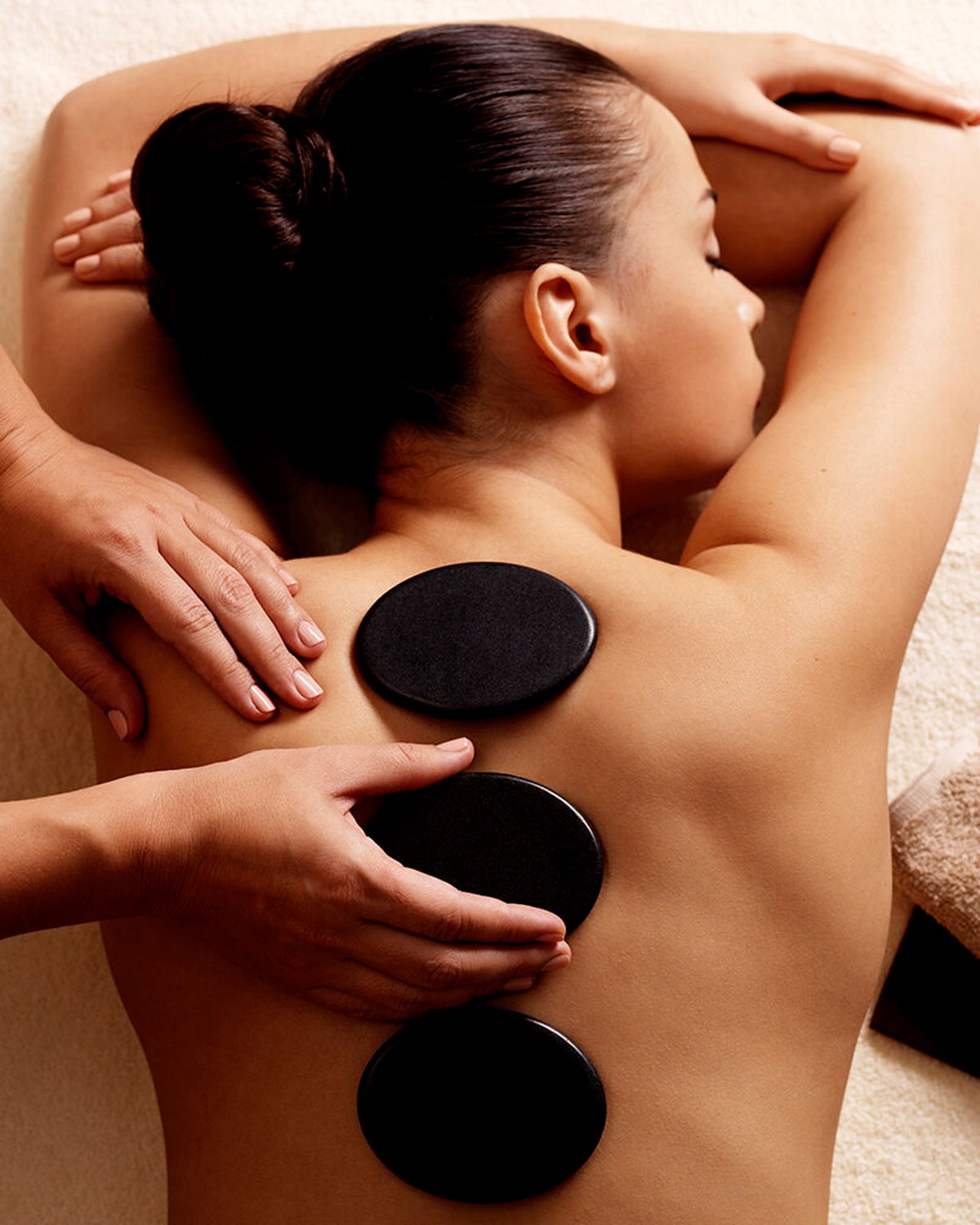 Woman relaxing face down on a soft towel with smooth black hot stones along her back while a therapist massages her shoulders during a luxury hot stone massage
