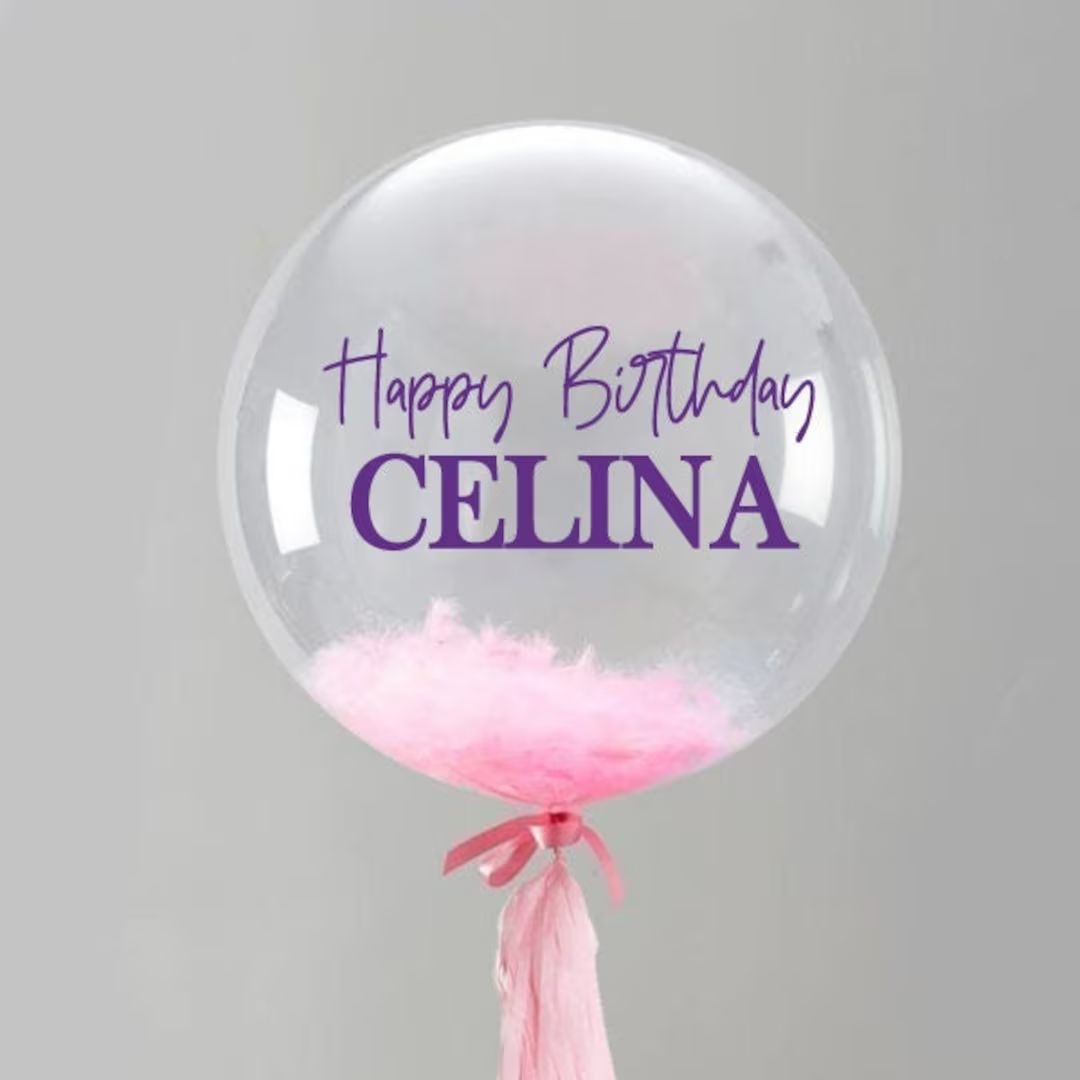 Personalized clear bubble balloon printed with Happy Birthday and a name, filled with pink feathers, with pink ribbon and tassel