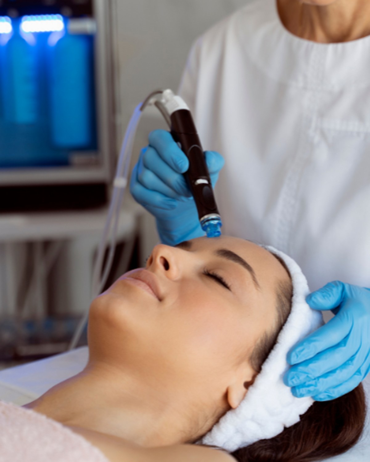 Woman relaxing with eyes closed and a white headband while a beautician in blue gloves treats her skin with a facial device during a Radiance & Rejuvenation facial